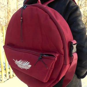Peut inclure: Un sac à dos bordeaux de forme circulaire. Il est doté d'une poche avant zippée avec un motif de plume blanche. Le sac à dos a une poignée supérieure, des bretelles réglables et une boucle latérale. Un cordon noir avec des perles pend de la fermeture éclair.
