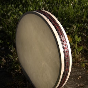 Op de afbeelding: Een ronde, crèmekleurige trommel met een decoratieve band met een rood en zwart patroon. De trommel staat tegen een achtergrond van groen gras, wat de textuur en het ontwerp benadrukt.