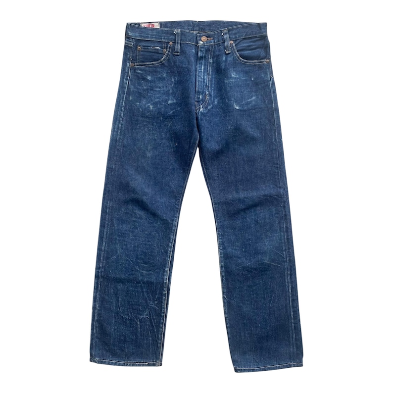 May include: A pair of dark blue denim jeans featuring a straight-leg cut, a classic five-pocket design, a button fly closure, and a visible red brand label on the interior waistband. The fabric shows subtle fading and whiskering patterns throughout.