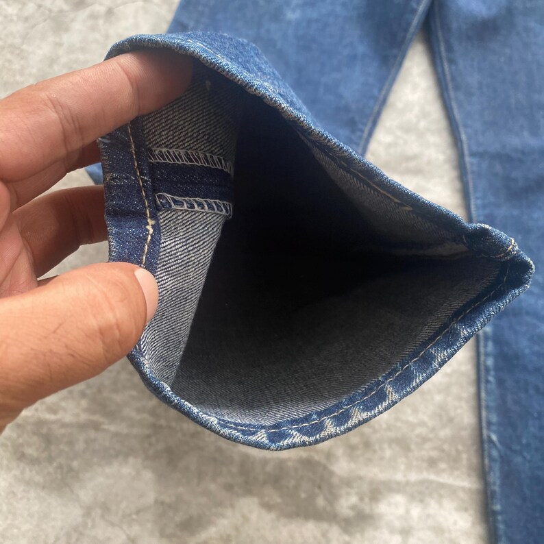 May include: A pair of dark blue denim jeans featuring a straight leg cut. The image shows the interior of the pant leg opening, revealing the dark blue exterior fabric, the lighter greyish-blue interior weave, and the white stitched hem detail.