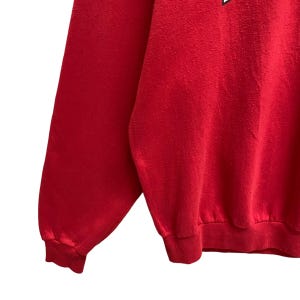 May include: A close-up of a red sweatshirt. The image shows the sleeve and part of the body of the sweatshirt. The fabric appears to be a soft, textured material. The cuffs and hem are ribbed.