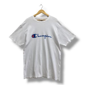 May include: White Champion t-shirt with the Champion logo in blue and red on the front. The short-sleeved shirt is hanging on a wooden hanger.