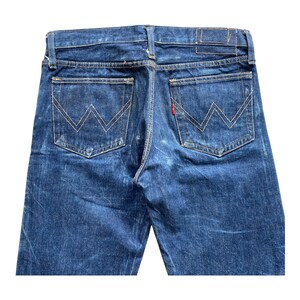 May include: Dark blue denim jeans featuring a classic five-pocket design with a high-rise waistband. The back pockets display a signature W-shaped stitching pattern in gold thread, with a small red tab attached to the right pocket edge.