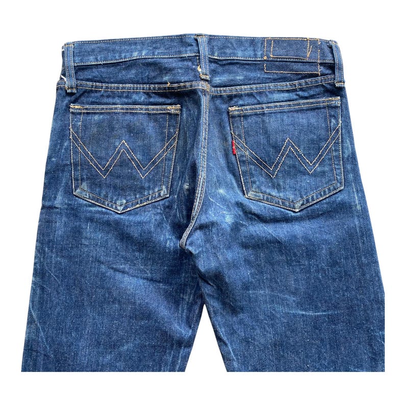 May include: Dark blue denim jeans featuring a classic five-pocket design with a high-rise waistband. The back pockets display a signature W-shaped stitching pattern in gold thread, with a small red tab attached to the right pocket edge.