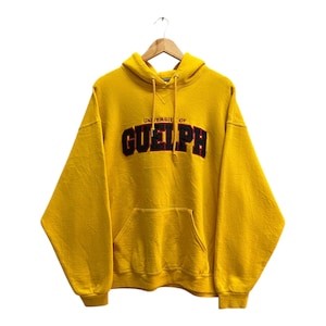 May include: Mustard yellow hoodie with a front pocket and drawstring hood. The text "UNIVERSITY OF GUELPH" is embroidered in black and red across the chest. The hoodie is hanging on a wooden hanger.