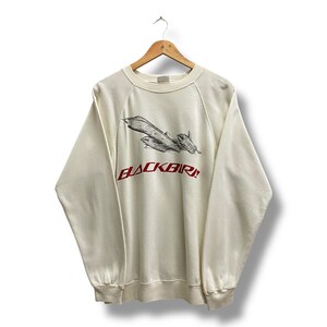 May include: Cream-colored sweatshirt with a black and red graphic of a Blackbird aircraft. The word "BLACKBIRD" is printed in red below the aircraft image. The sweatshirt is hanging on a wooden hanger.