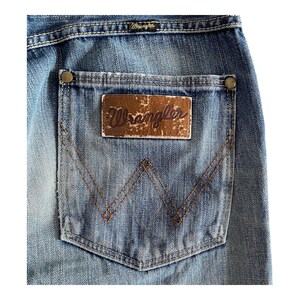 Pu&ograve; includere: Un paio di jeans blu denim con una tasca posteriore che presenta una toppa in pelle marrone con la parola "Wrangler" in cuciture viola. La tasca ha una distintiva "W" cucita in filo marrone.