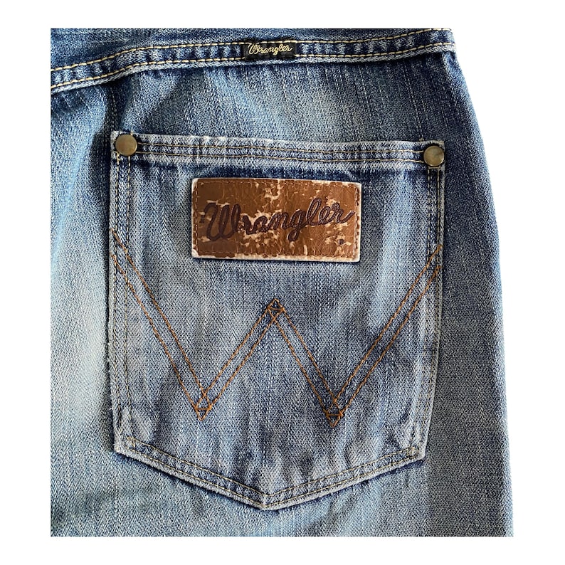 Pu&ograve; includere: Un paio di jeans blu denim con una tasca posteriore che presenta una toppa in pelle marrone con la parola "Wrangler" in cuciture viola. La tasca ha una distintiva "W" cucita in filo marrone.