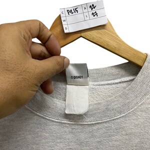 May include: A light gray sweatshirt with a crew neck, held by a wooden hanger. The tag reads "&copy; DISNEY". A white tag is attached to the sweatshirt. A price tag is attached to the hanger with the numbers 22 and 27.