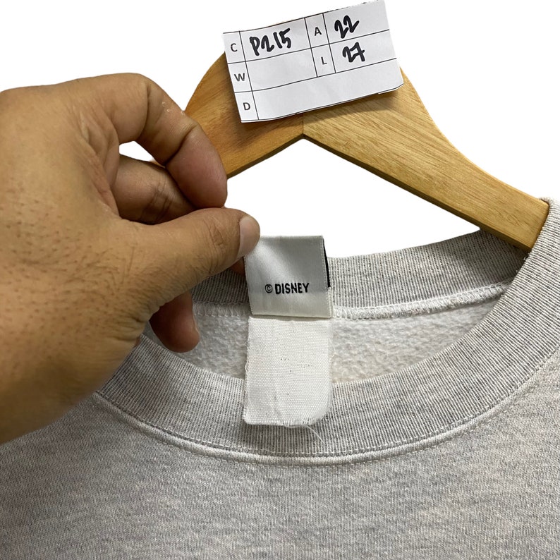 May include: A light gray sweatshirt with a crew neck, held by a wooden hanger. The tag reads "&copy; DISNEY". A white tag is attached to the sweatshirt. A price tag is attached to the hanger with the numbers 22 and 27.