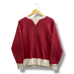 May include: A red sweatshirt with a gray neckline, cuffs, and hem. The long-sleeved top is hanging on a wooden hanger against a white background. The neckline has a V-shape.