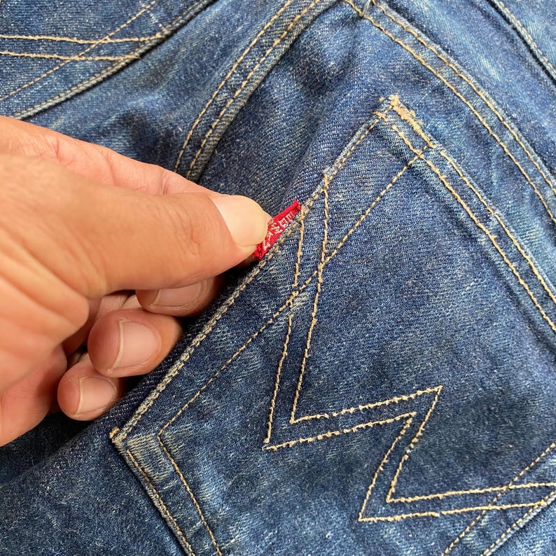May include: Dark blue denim jeans featuring a back pocket with a distinctive W-shaped stitching pattern in gold thread. A small red fabric tab is attached to the side of the pocket, showcasing the classic construction of these durable trousers.