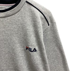 Puede incluir: Sudadera gris de cuello redondo con el logotipo de FILA bordado en rojo y azul marino. La sudadera tiene un cuello a rayas blanco y negro y una raya azul marino en el hombro. La tela parece ser un material suave y c&oacute;modo.