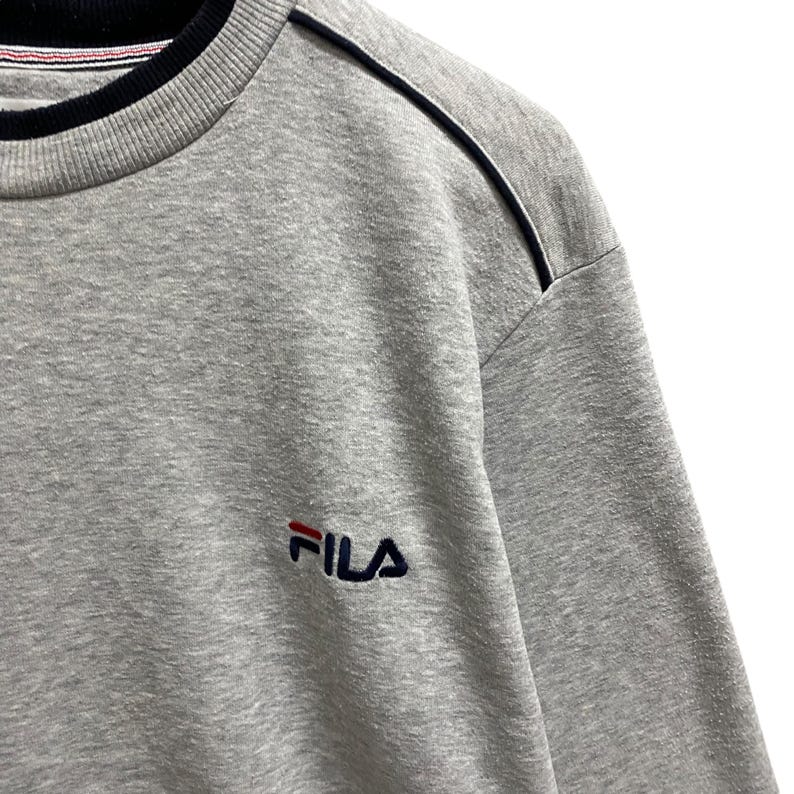 Puede incluir: Sudadera gris de cuello redondo con el logotipo de FILA bordado en rojo y azul marino. La sudadera tiene un cuello a rayas blanco y negro y una raya azul marino en el hombro. La tela parece ser un material suave y c&oacute;modo.