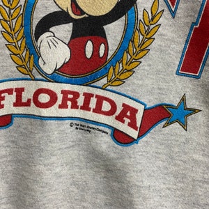 May include: A heather gray sweatshirt featuring a graphic of Mickey Mouse inside a blue circle framed by gold laurel leaves. Below, a red banner with the text FLORIDA is displayed, accompanied by a blue star. The text The Walt Disney Company by Sherry Mfg is printed below the banner.