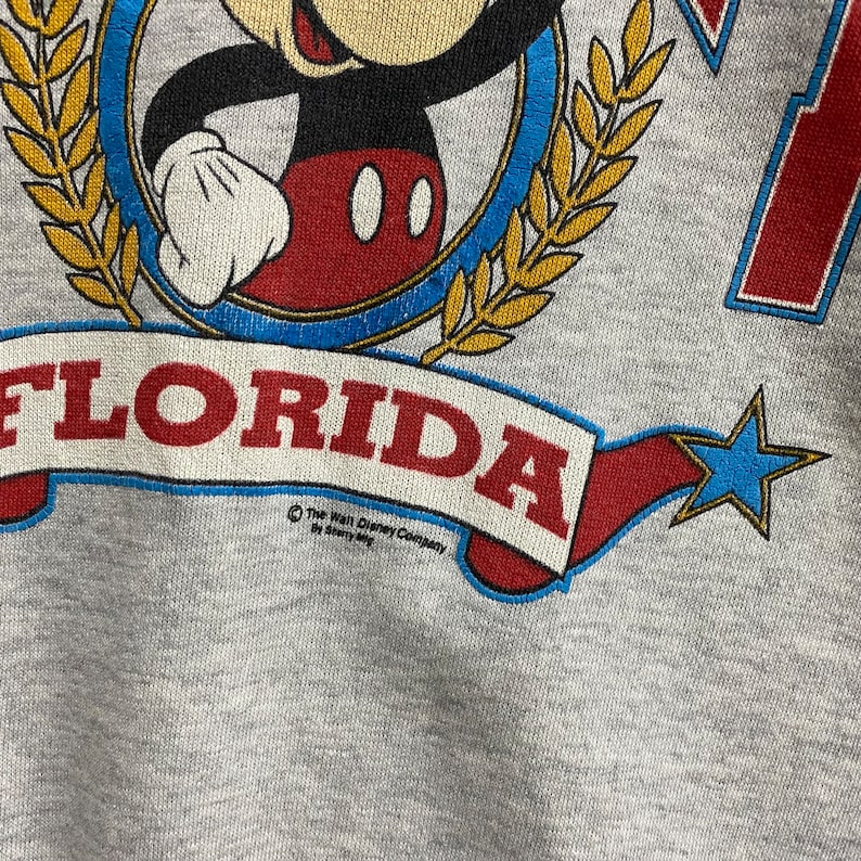 May include: A heather gray sweatshirt featuring a graphic of Mickey Mouse inside a blue circle framed by gold laurel leaves. Below, a red banner with the text FLORIDA is displayed, accompanied by a blue star. The text The Walt Disney Company by Sherry Mfg is printed below the banner.