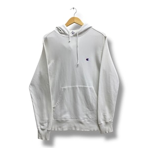 Puede incluir: Una sudadera con capucha Champion blanca con capucha con cordón y bolsillo tipo canguro. La sudadera con capucha presenta el logotipo de Champion en un pequeño parche bordado en el pecho izquierdo. La sudadera está colgada en una percha de madera.