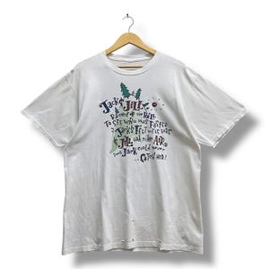 Puede incluir: Camiseta blanca con un diseño gráfico que presenta el texto "Jack & Jill" y la frase "Raced up the hill." El diseño incluye ilustraciones de árboles, un sol y las palabras "Jack's feet were bare."