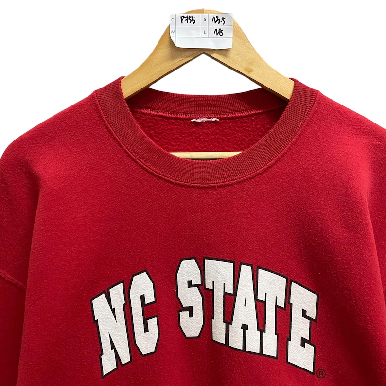 May include: A red sweatshirt with the words "NC STATE" in white block letters. The sweatshirt is hanging on a wooden hanger. The tag on the hanger has the numbers "13.5" and "16".