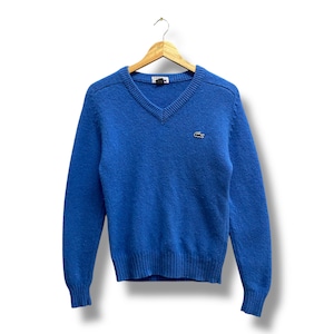 May include: A blue V-neck sweater with long sleeves, hanging on a wooden hanger. The sweater has a ribbed hem and cuffs, and a small green crocodile logo on the chest.