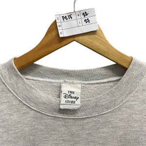 May include: A light gray sweatshirt with a crew neck, hanging on a wooden hanger. The tag reads "THE DISNEY STORE". A white tag with the numbers "22" and "27" is attached to the hanger.