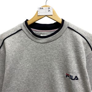 Puede incluir: Una sudadera gris de FILA con cuello y ribetes azul marino. La sudadera tiene un peque&ntilde;o logotipo de FILA en el pecho y est&aacute; colgada de una percha de madera. El cuello tiene rayas rojas y blancas.