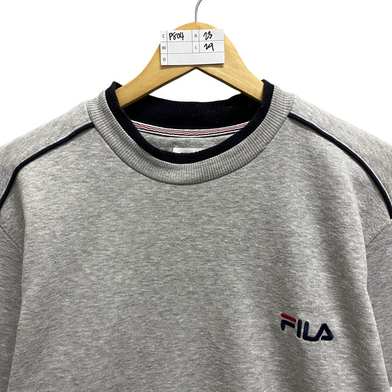 Puede incluir: Una sudadera gris de FILA con cuello y ribetes azul marino. La sudadera tiene un peque&ntilde;o logotipo de FILA en el pecho y est&aacute; colgada de una percha de madera. El cuello tiene rayas rojas y blancas.