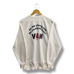Pode incluir: Camisola branca com o texto "for the young and the young-at-heart" em preto e azul marinho, com "VAN" em vermelho, azul marinho e preto. A camisola está pendurada num cabide de madeira.