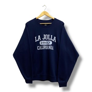 May include: Navy blue sweatshirt with the text "LA JOLLA 1887 CALIFORNIA" in white. The sweatshirt is hanging on a wooden hanger. The design is centered on the front of the garment.