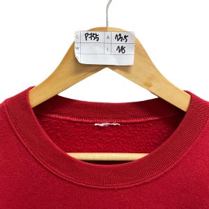 May include: A red crew neck sweatshirt hanging on a wooden hanger. A white tag with black handwritten numbers and letters is attached to the hanger. The sweatshirt is made of a soft material.