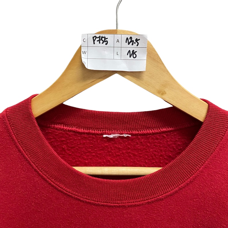 May include: A red crew neck sweatshirt hanging on a wooden hanger. A white tag with black handwritten numbers and letters is attached to the hanger. The sweatshirt is made of a soft material.