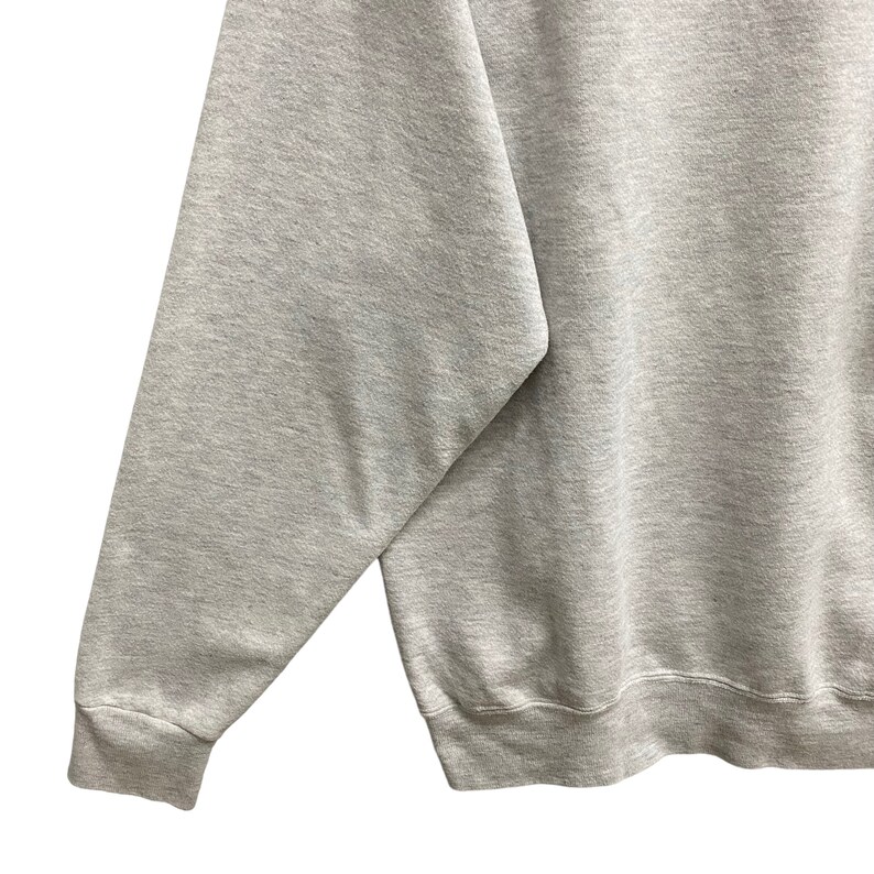 May include: A light gray sweatshirt with a crew neck and long sleeves. The image shows a close-up of the sleeve and part of the body of the sweatshirt. The fabric appears to be a soft, comfortable material.