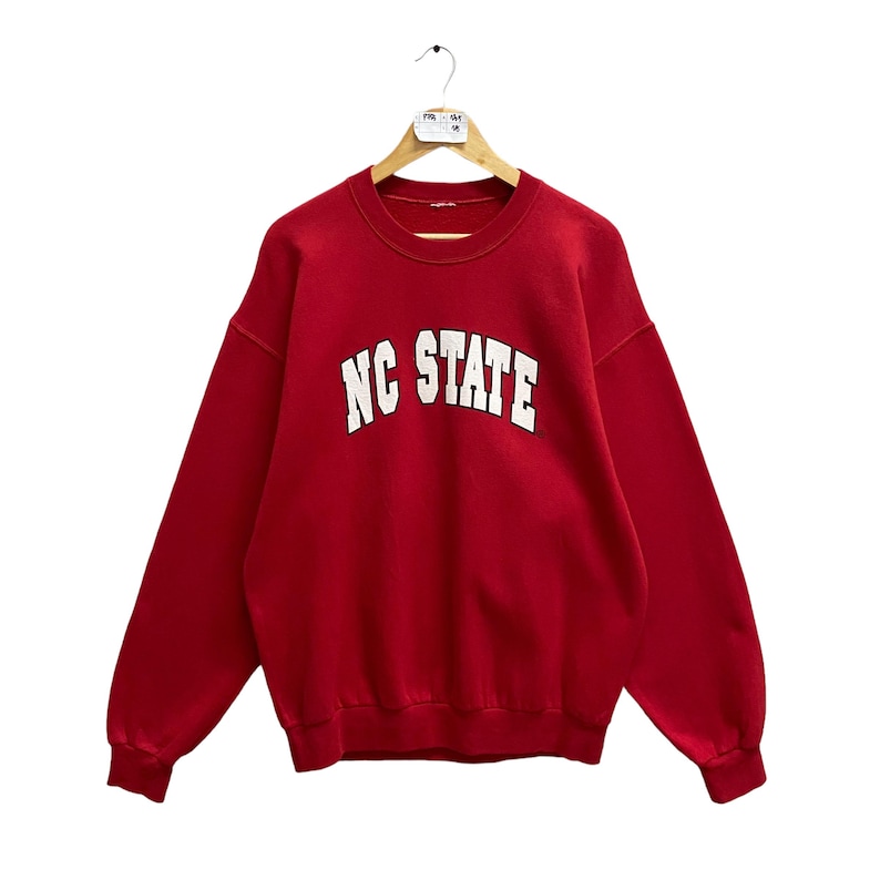 May include: A red sweatshirt with the text "NC STATE" in white block letters. The sweatshirt has long sleeves and a crew neck. The garment is hanging on a wooden hanger.