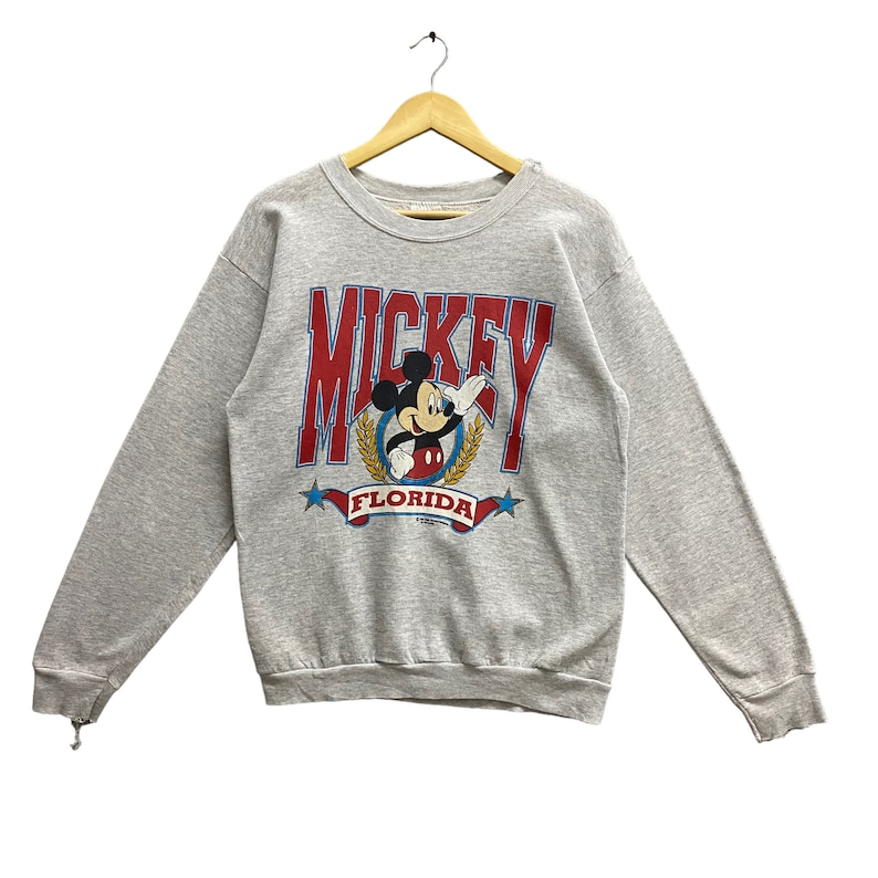 May include: A heather gray crewneck sweatshirt featuring a graphic of Mickey Mouse centered within a gold laurel wreath. The word MICKEY is printed in large red letters above, with FLORIDA written on a red banner below, flanked by two blue stars.