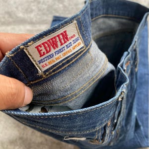 May include: Dark blue denim jeans featuring a rectangular white fabric label stitched to the interior waistband. The label displays the red text EDWIN and blue text reading WESTERN FINEST BLUE JEANS U.S. REG. PAT. OFFICE APPLIED.