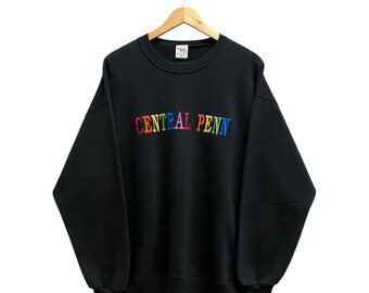 Pode incluir: Camisola preta de gola redonda com o texto bordado "CENTRAL PENN" em cores do arco-íris. A camisola está pendurada num cabide de madeira sobre um fundo branco. Tem mangas compridas e decote canelado.