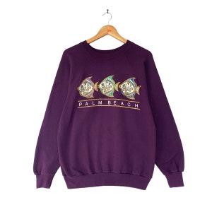 May include: A purple sweatshirt with a crew neck and long sleeves. The front features three stylized fish in shades of purple, teal, and gold, with the text "PALM BEACH" below them in gold lettering.