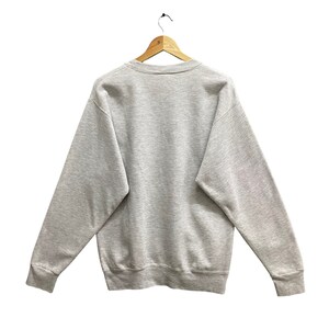 May include: A heather gray sweatshirt with a crew neck and long sleeves, hanging on a wooden hanger. The sweatshirt is a solid color, with ribbed cuffs and hem. The back of the sweatshirt is visible.
