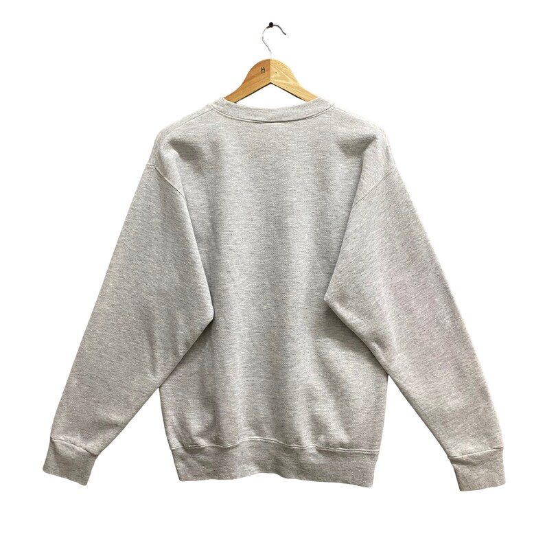 May include: A heather gray sweatshirt with a crew neck and long sleeves, hanging on a wooden hanger. The sweatshirt is a solid color, with ribbed cuffs and hem. The back of the sweatshirt is visible.