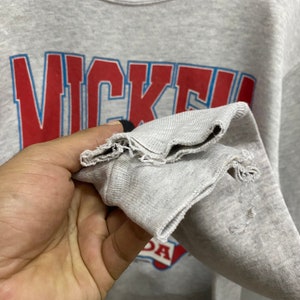 May include: A light gray crewneck sweatshirt featuring the red and blue text "NICKE" printed on the front. The sleeve cuff shows visible fraying and wear along the hemline, with additional distressed fabric texture near the wrist area.