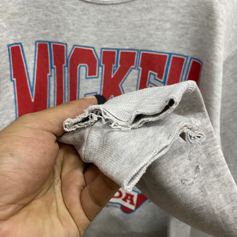 May include: A light gray crewneck sweatshirt featuring the red and blue text "NICKE" printed on the front. The sleeve cuff shows visible fraying and wear along the hemline, with additional distressed fabric texture near the wrist area.