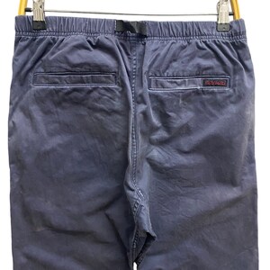 May include: Navy blue pants featuring an elastic waistband with an integrated black nylon webbing belt, two rear welt pockets, and a small red Gramicci brand label stitched onto the right pocket edge, all suspended from a yellow plastic hanger.