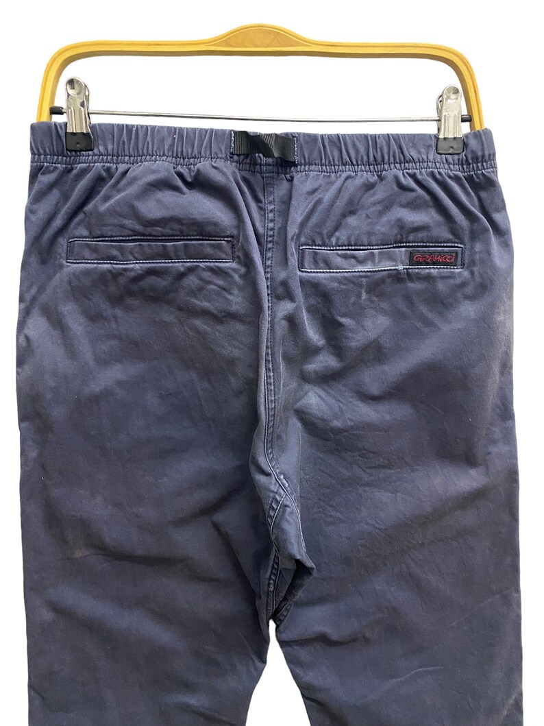 May include: Navy blue pants featuring an elastic waistband with an integrated black nylon webbing belt, two rear welt pockets, and a small red Gramicci brand label stitched onto the right pocket edge, all suspended from a yellow plastic hanger.