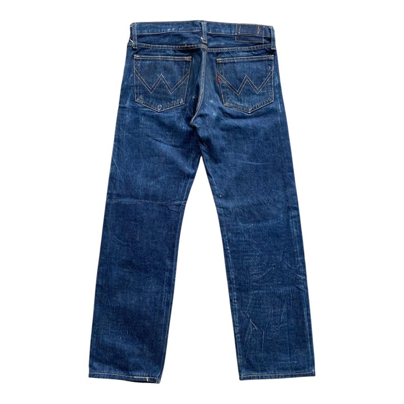 May include: A pair of dark blue denim jeans featuring a straight leg cut, a waistband with belt loops, and two back pockets. Each back pocket is detailed with a distinct white stitched W-shaped design and a small red tab on the right pocket.