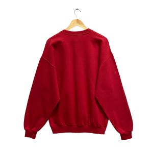 May include: A solid red sweatshirt hangs from a wooden hanger against a white background. The crew neck sweatshirt has long sleeves and ribbed cuffs. The back of the sweatshirt is visible, showcasing its simple design and color.