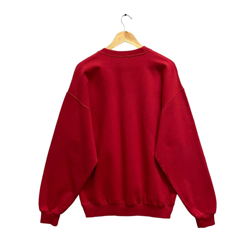 May include: A solid red sweatshirt hangs from a wooden hanger against a white background. The crew neck sweatshirt has long sleeves and ribbed cuffs. The back of the sweatshirt is visible, showcasing its simple design and color.