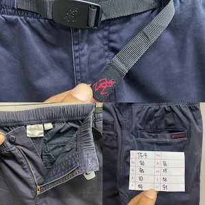 May include: Navy blue pants featuring an integrated black nylon belt with a plastic buckle, an elastic waistband, a metal zipper fly, and a Gramicci brand label. A white paper tag displays handwritten measurements in inches for waist, hip, and length.