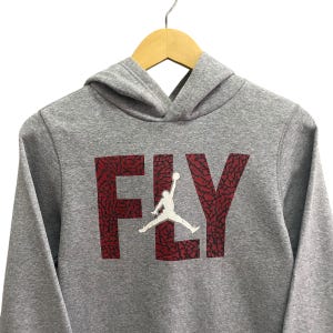 Puede incluir: Una sudadera con capucha gris con la palabra "FLY" en rojo, con una silueta blanca de un jugador de baloncesto. La sudadera con capucha est&aacute; colgada en una percha de madera.