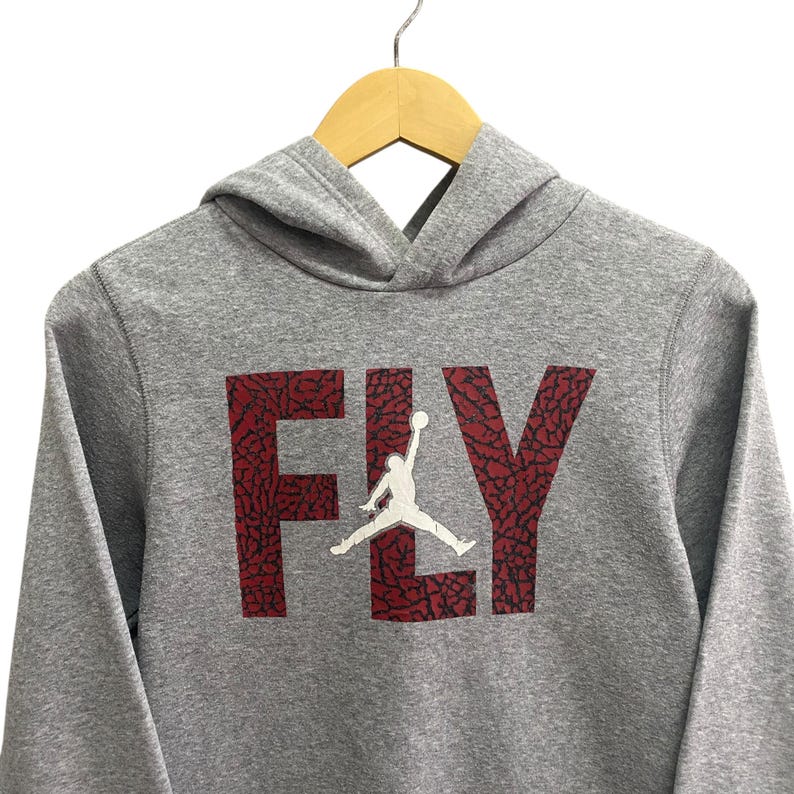 Puede incluir: Una sudadera con capucha gris con la palabra "FLY" en rojo, con una silueta blanca de un jugador de baloncesto. La sudadera con capucha est&aacute; colgada en una percha de madera.