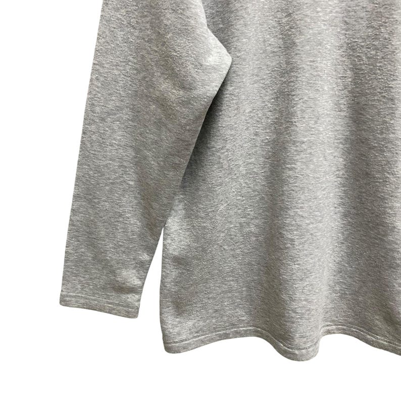 Puede incluir: Primer plano de una camiseta gris de manga larga. La camiseta est&aacute; hecha de una tela suave y texturizada. La manga es visible en el lado izquierdo de la imagen, con el cuerpo de la camiseta a la derecha. El dobladillo de la camiseta es visible en la parte inferior.